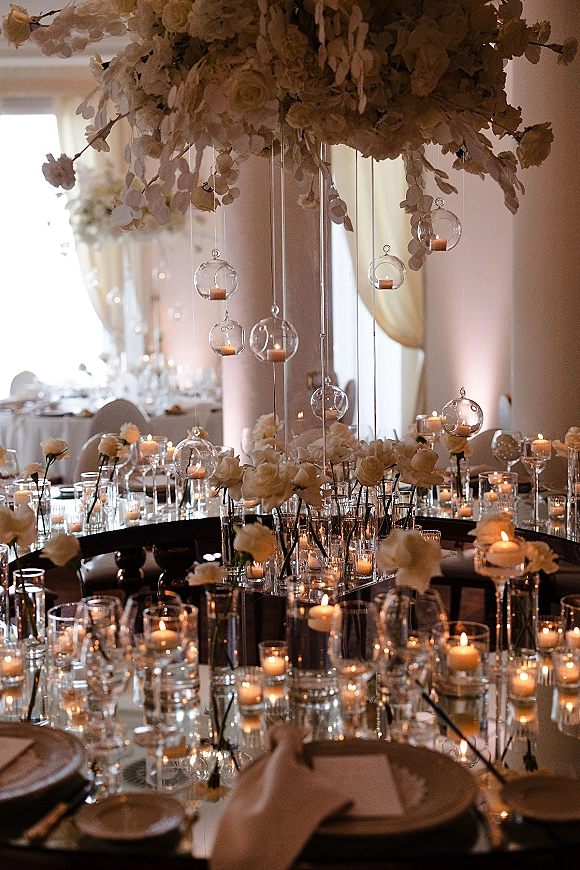 Reception tablescape with candlelit wedding table styling, white rose centerpieces and hanging glass votives in a draped ballroom with columns