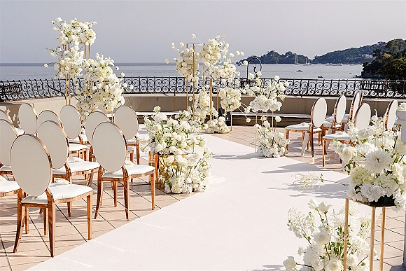 Ceremony setup with a white aisle runner and floral arrangements on gold stands, set on a terrace patio overlooking the ocean view