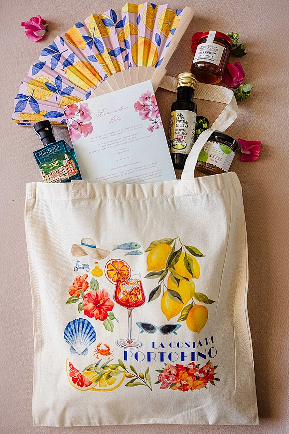 Wedding welcome bags in canvas tote with floral invitation and hand fan, plus mini liquor, olive oil, and honey favors on neutral fabric