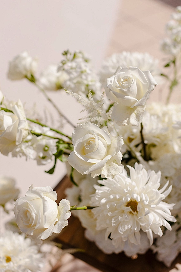 White wedding flowers with white roses and chrysanthemums arranged with greenery stems on a beige surface in soft sunlight and shadows