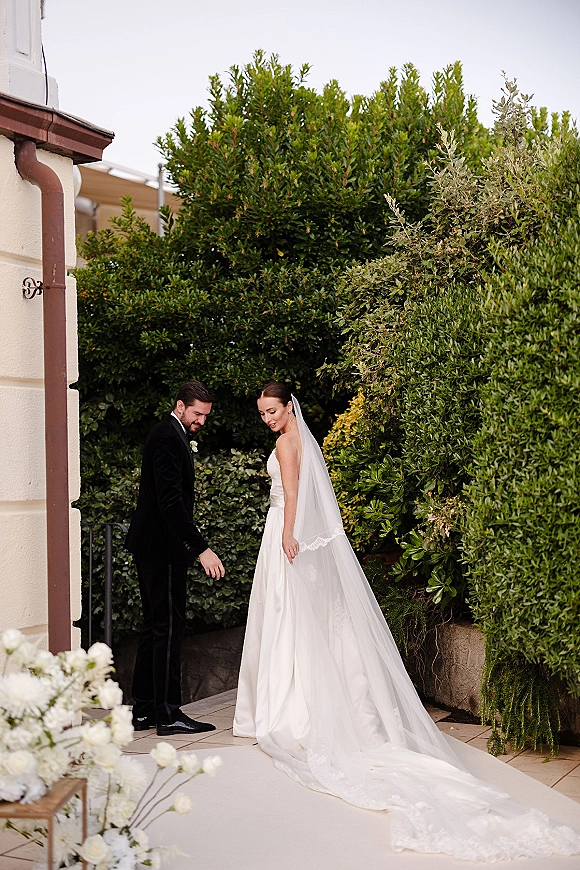 Couple portrait of bride in a long veil and strapless gown with groom in black tuxedo beside a white floral arrangement on a garden patio