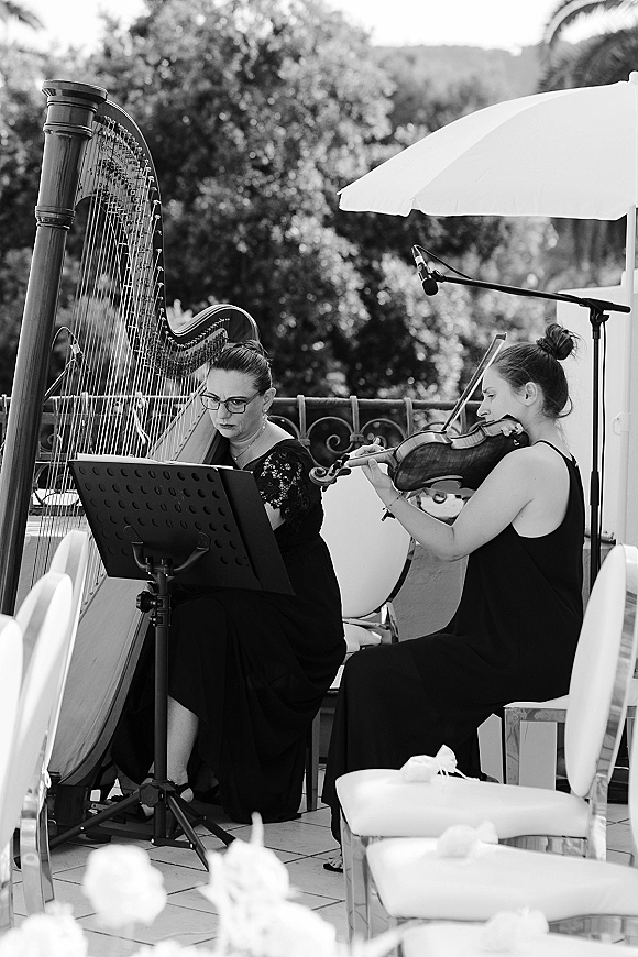 Wedding musicians perform harp and violin duo beside a sheet music stand on an outdoor patio with chairs, trees, and distant hills