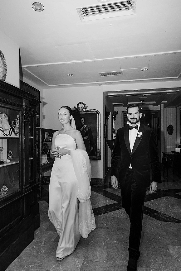 Bride and groom in a black and white wedding photo walking hand in hand through an indoor hallway, her long veil trailing beside his tuxedo