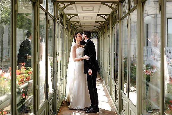 Wedding kiss portrait of bride and groom kiss in a glass corridor with green frames, her veil flowing as they embrace on wood floors