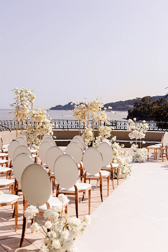 Ceremony setup with outdoor ceremony seating and a white aisle runner, gold stands and white florals on a terrace overlooking the ocean coastline