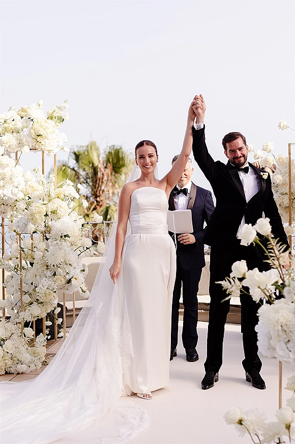 Wedding recessional with bride and groom recessional raising hands, walking past white florals on gold frames along an outdoor terrace aisle runner