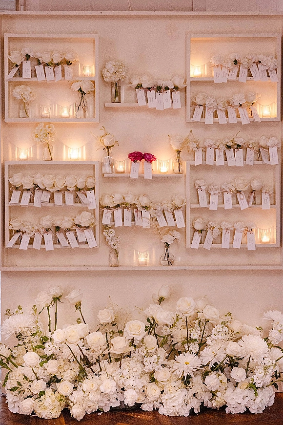Escort card display with wedding escort cards tied with white ribbon among white and blush roses on wall shelves in warm lighting