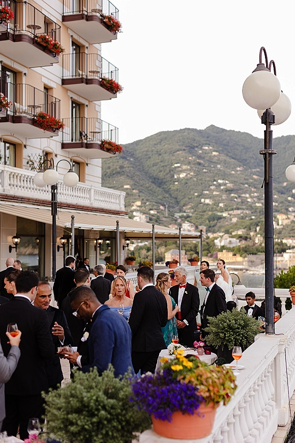 Wedding cocktail hour with wedding guests mingling on a hotel terrace, holding Aperol spritz glasses amid potted florals and mountain waterfront views