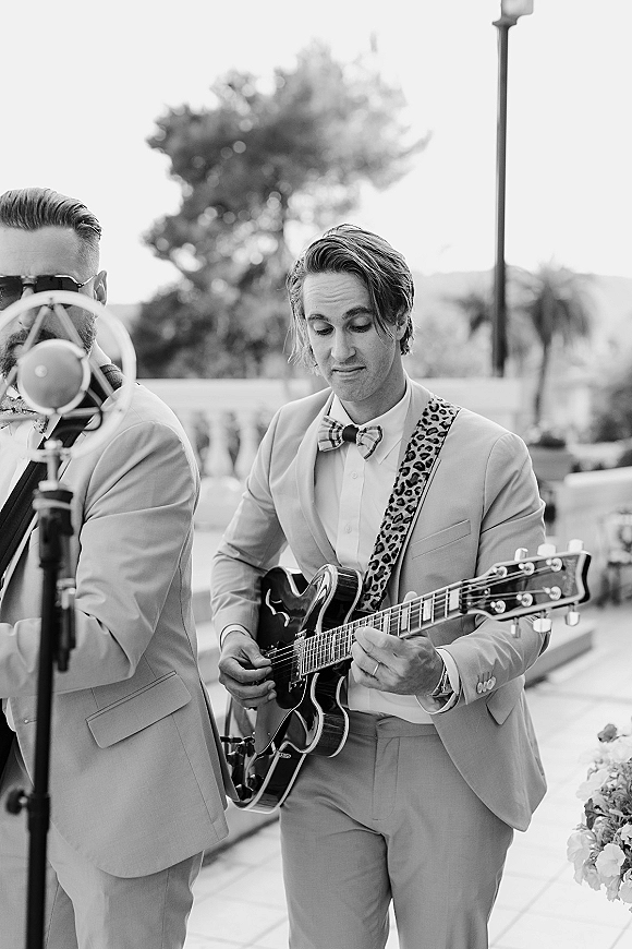 Wedding band of suited musicians playing electric guitar and singing into a microphone on an outdoor terrace with mountains behind