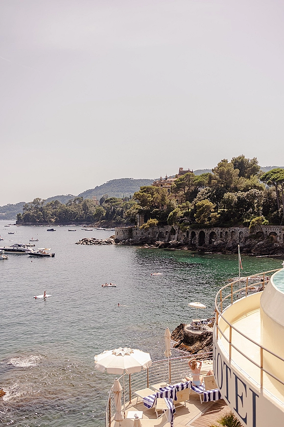 Coastal landscape from a seaside terrace view, with boats on calm turquoise water, beach umbrellas and loungers above a rocky cove
