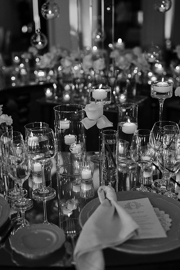 Reception tablescape with candlelit wedding table glow, floating candles in clear glass cylinders, white roses, and menu cards in a dim room