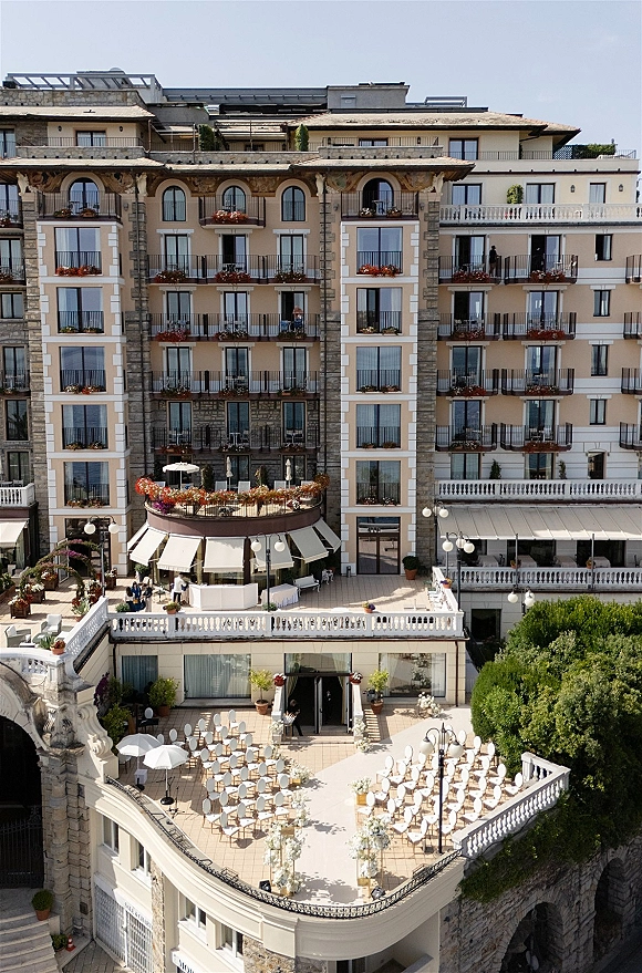 Ceremony setup for an outdoor wedding ceremony with white chairs and floral aisle arrangements on a stone hotel terrace under umbrellas