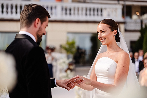 Ring exchange as bride in a strapless wedding dress places a wedding ring on groom in tuxedo during an outdoor ceremony with guests