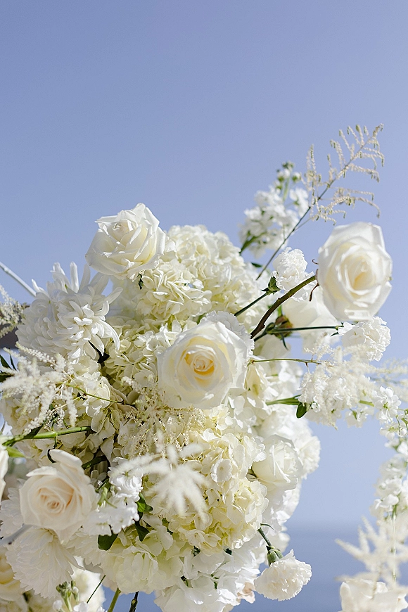 Wedding bouquet of white roses and hydrangea with greenery accents held up against a clear blue sky, featuring soft all-white blooms