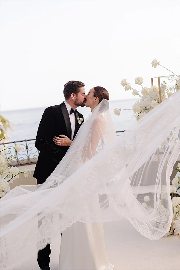 Wedding kiss portrait of bride and groom kissing under a white floral arch, cathedral veil flowing on an ocean-view terrace