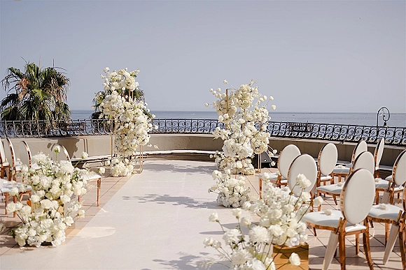 Ceremony setup with white florals on gold stands lining a white aisle runner, facing an oceanfront terrace with palm trees and railing