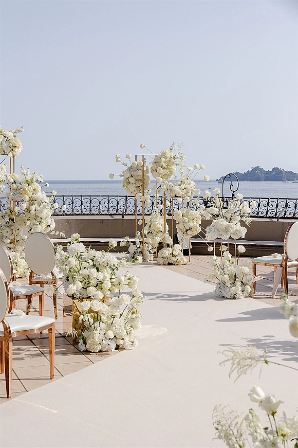 Ceremony setup for outdoor wedding ceremony with white florals and gold frame stands lining a white aisle runner on an oceanview terrace