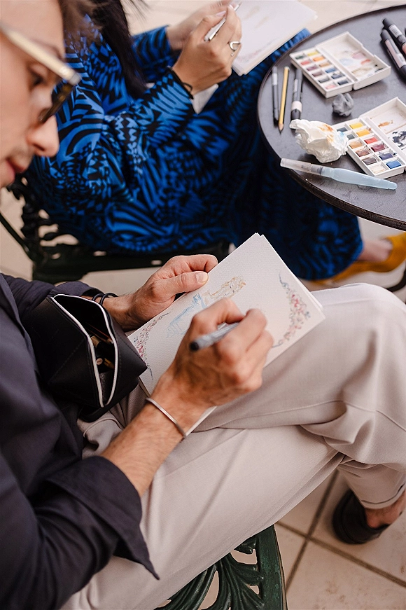 Wedding live painting setup with watercolor paint set on a table, brushes and pens beside sketchbook pages, chairs on tiled floor behind
