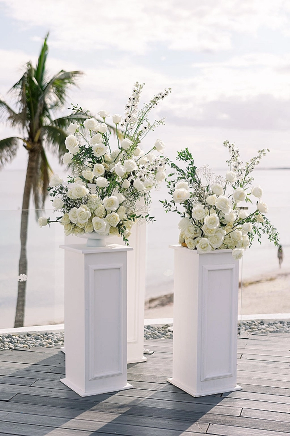 Ceremony floral pillars with white rose ceremony flowers on tall pedestals, framing a beach deck aisle with ocean and palm backdrop