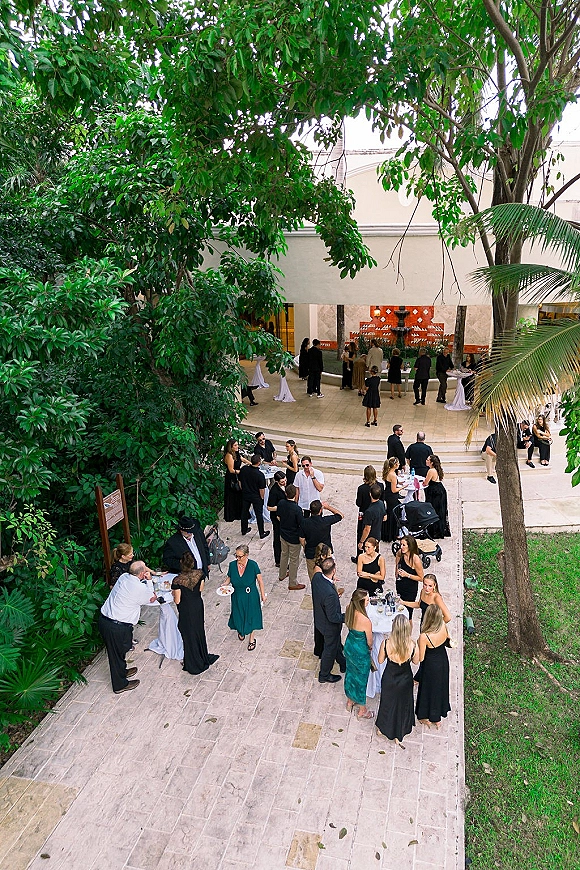 Wedding cocktail hour with high-top tables in white linens, guests mingling with drinks around a courtyard fountain under trees