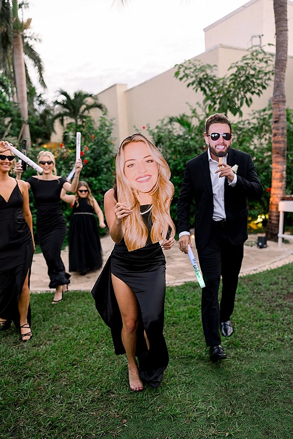 Wedding exit with bridesmaids in black dresses and groom in sunglasses waving glow sticks and a big face cutout on a palm-lined walkway