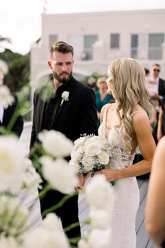 Ceremony moment as bride and groom walk down the aisle, she holds white rose bouquet in lace gown beside guests on terrace