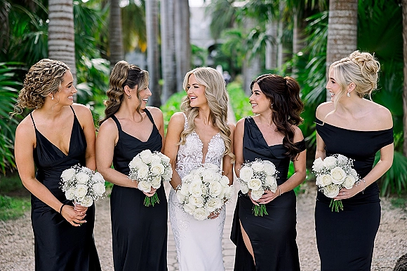Bridal party portrait of bride with bridesmaids holding white rose bouquets, bride in lace gown, on a palm-lined tropical garden path outdoors