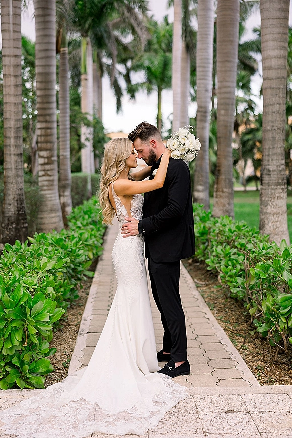 Couple portrait of bride and groom embrace with white rose bouquet, lace open-back gown and black tuxedo on palm-lined walkway