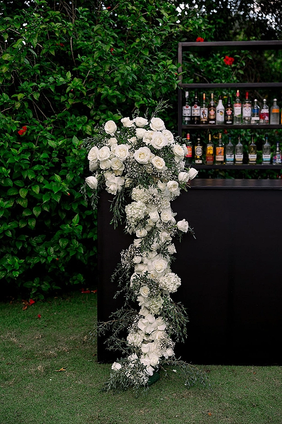 Wedding bar setup with liquor bottles on a black counter, styled as an outdoor wedding bar with white roses and eucalyptus on a lawn