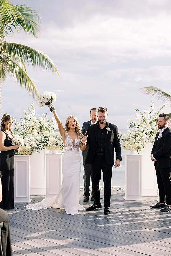 Recessional moment as newlyweds walk down the aisle, bride holding bouquet up on an oceanfront deck with palm trees and sky behind