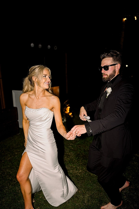 Reception dance as bride in a strapless satin wedding dress spins with groom in sunglasses under string lights on an outdoor lawn at night
