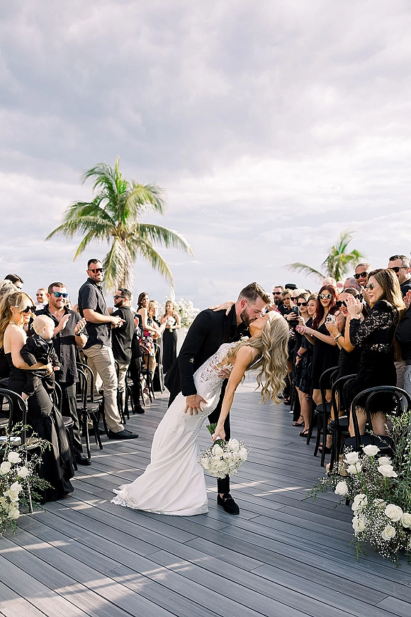Wedding kiss as the bride dips the groom in the aisle, holding a white rose bouquet on an outdoor deck with palm trees and guests cheering