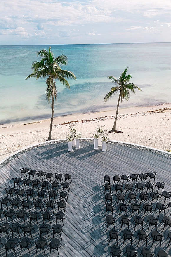 Beach ceremony setup with oceanfront wedding ceremony seating in curved rows of black chairs around floral pillars on a wooden deck by the sea