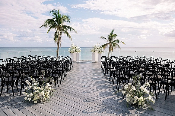 Beach wedding ceremony setup with an oceanfront ceremony aisle lined with white florals and black chairs on a wooden deck by palm trees