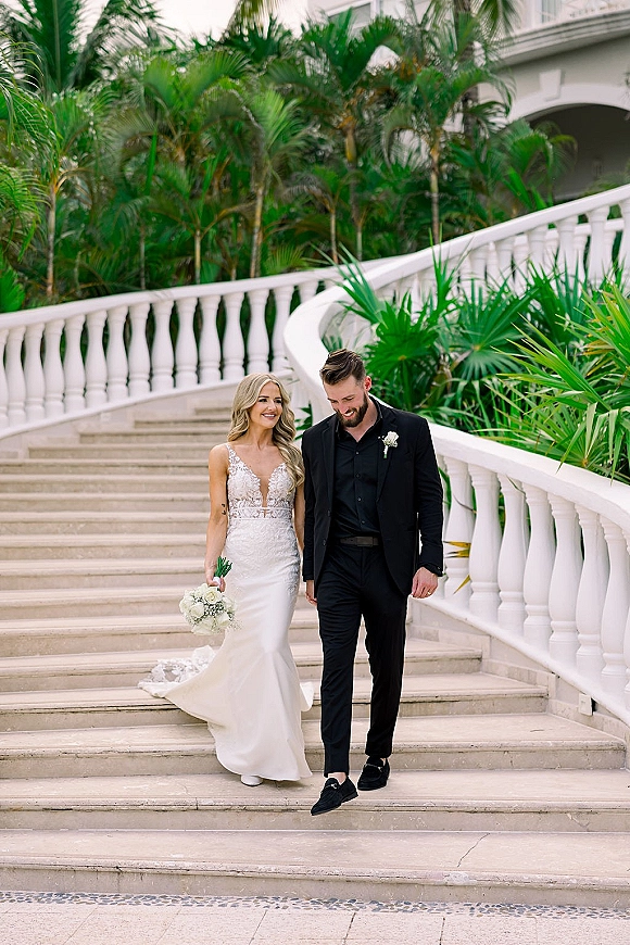 Couple portrait of newlyweds on stairs, bride in lace wedding dress holding bouquet beside groom in black suit by white balustrade