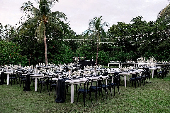 Outdoor reception decor with long banquet tables set in black and white, white floral centerpieces under string lights on a tropical lawn