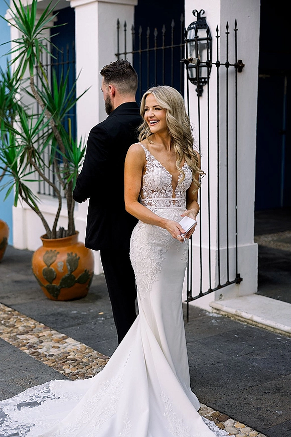 First look moment as bride and groom stand back to back by a wrought iron gate, bride in lace dress holding a handwritten note outdoors