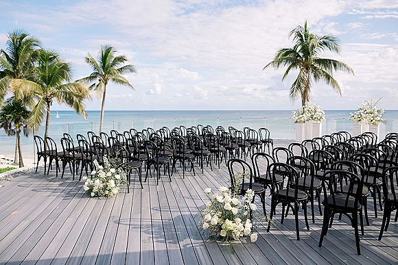 Beach ceremony setup with oceanfront wedding ceremony seating, black bentwood chairs and white florals on a wooden deck by the sea