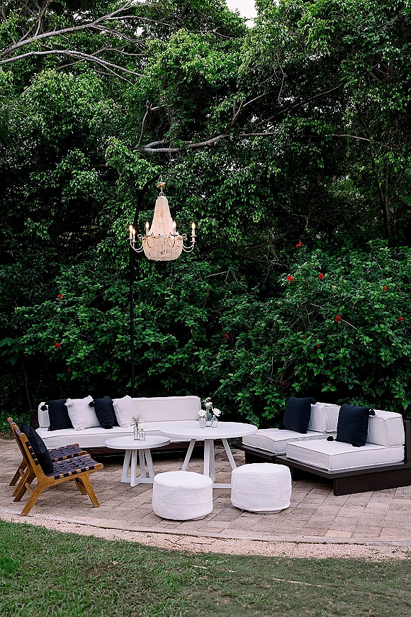 Outdoor lounge seating with a white sectional, black throw pillows, and coffee tables with small florals on a stone patio in a garden setting