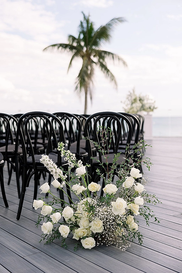 Ceremony aisle decor with outdoor ceremony seating, black chairs and a white rose and greenery ground arrangement on a wood deck by the ocean view