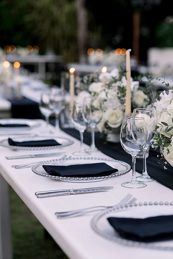 Wedding tablescape with black and white wedding table styling, white rose centerpiece and greenery garland with taper candles under bistro lights outdoors