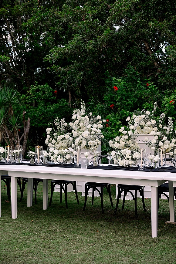 Reception tablescape with an outdoor reception table featuring white rose centerpieces, greenery garland, and candlelit glass vases on a garden lawn