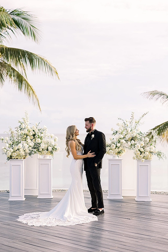 Couple portrait of bride and groom on a wooden oceanfront deck, her long train flowing beside white floral pedestals and palm trees