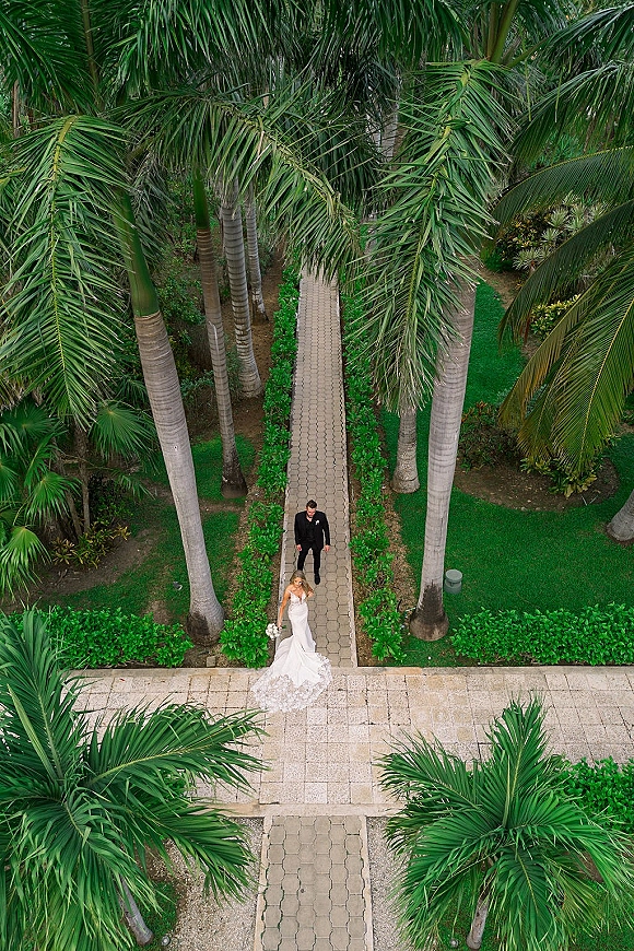 Couple portrait of bride and groom walking down a palm-lined garden walkway, bride holding a bouquet with lace train trailing behind