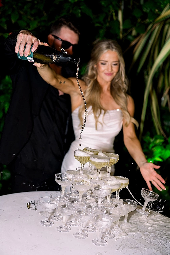 Champagne tower as newlyweds pour from a bottle into coupe glasses, bride in dress and groom in suit with sunglasses amid tropical greenery at night