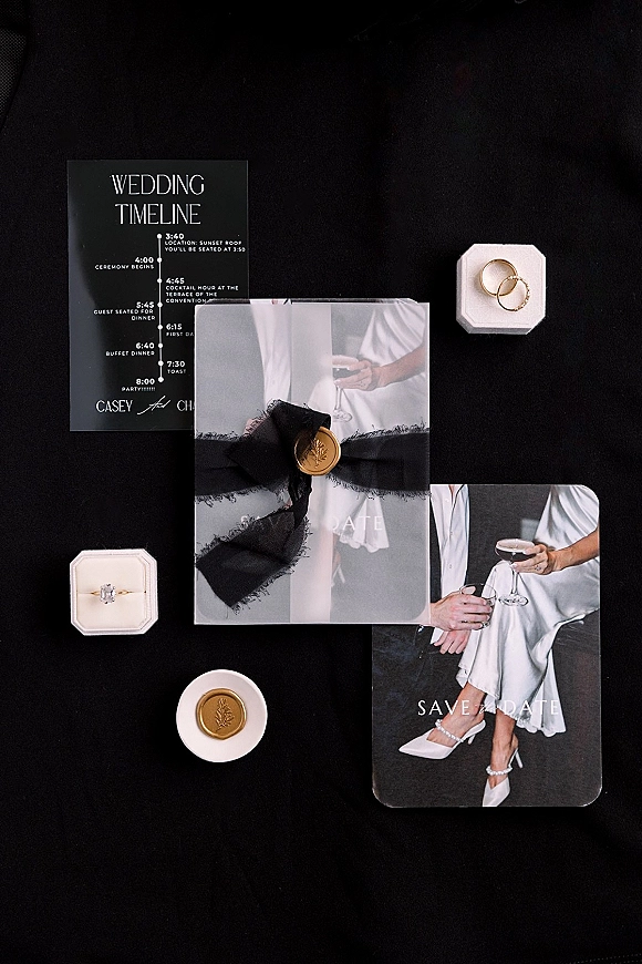 Wedding invitation suite with black wedding invitations, save the date and timeline cards, ribbon wrap, wax seal, and rings on black fabric backdrop