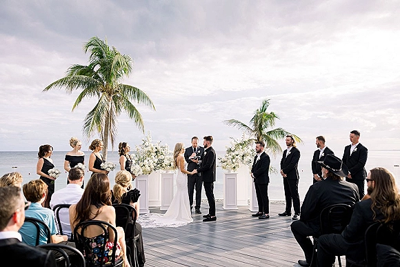 Wedding ceremony at a white floral arch with bride and groom on a wood deck, ocean and palm trees behind, bridesmaids and guests seated