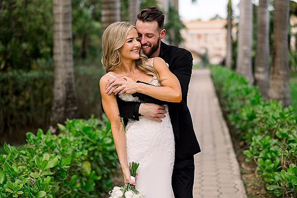 Couple portrait of bride and groom embrace on a garden pathway, groom hugging bride from behind as she holds a white rose bouquet by palm trees