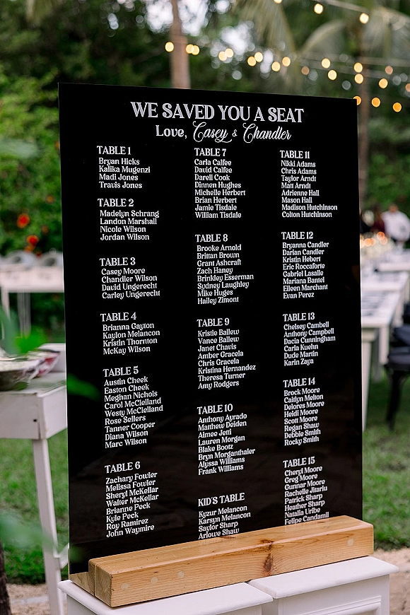 Wedding seating chart sign in black acrylic with white lettering on a wooden base at an outdoor reception with string lights and guests