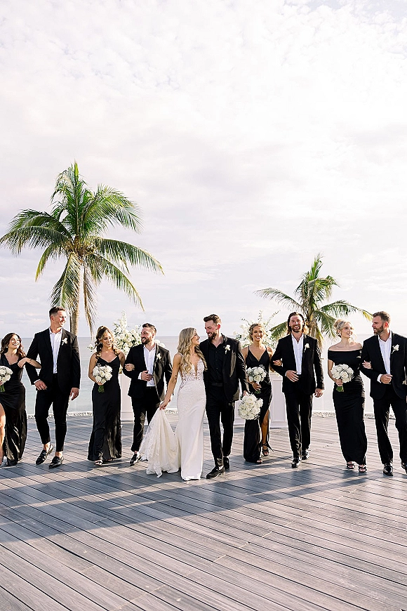 Wedding party portrait of bride and groom with bridesmaids and groomsmen, black suits and white rose bouquets on an oceanfront deck with palms